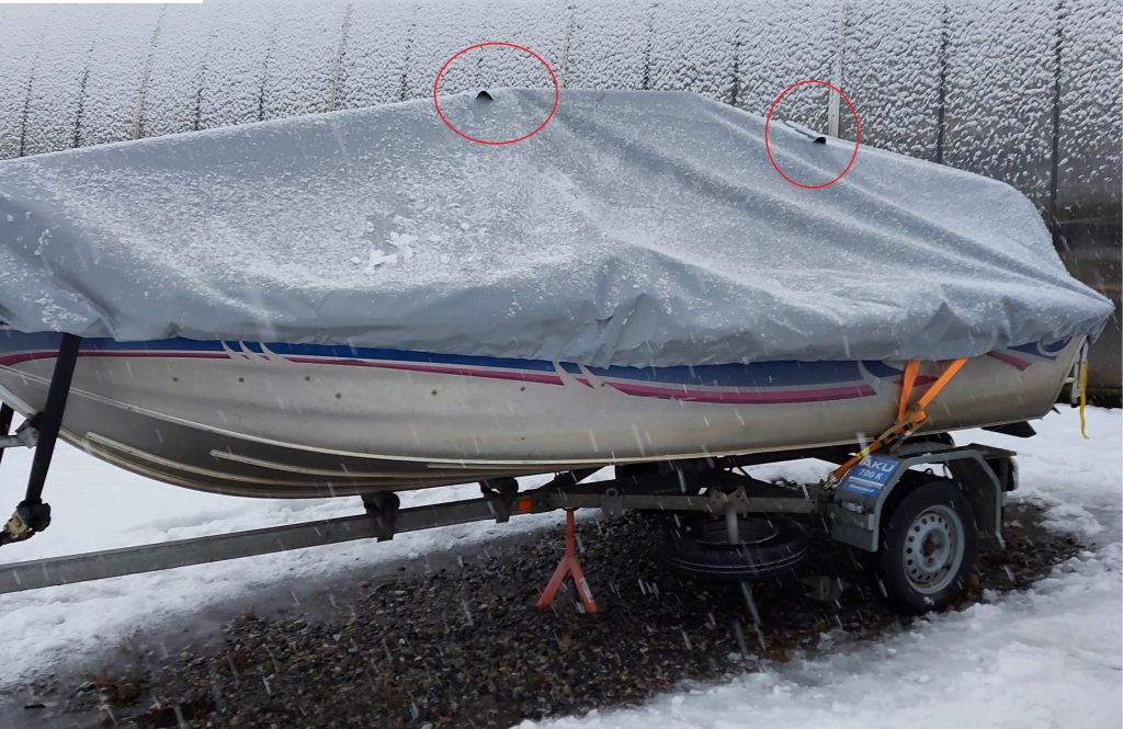 Boat on a trailer under a grey PVC cover in falling snow, with the two ventilation openings circled